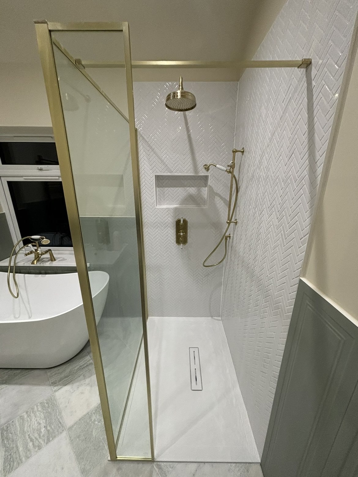 Walk-in shower with brushed brass framed screen, rainfall head, handset and recessed shelf niche in white herringbone tiling