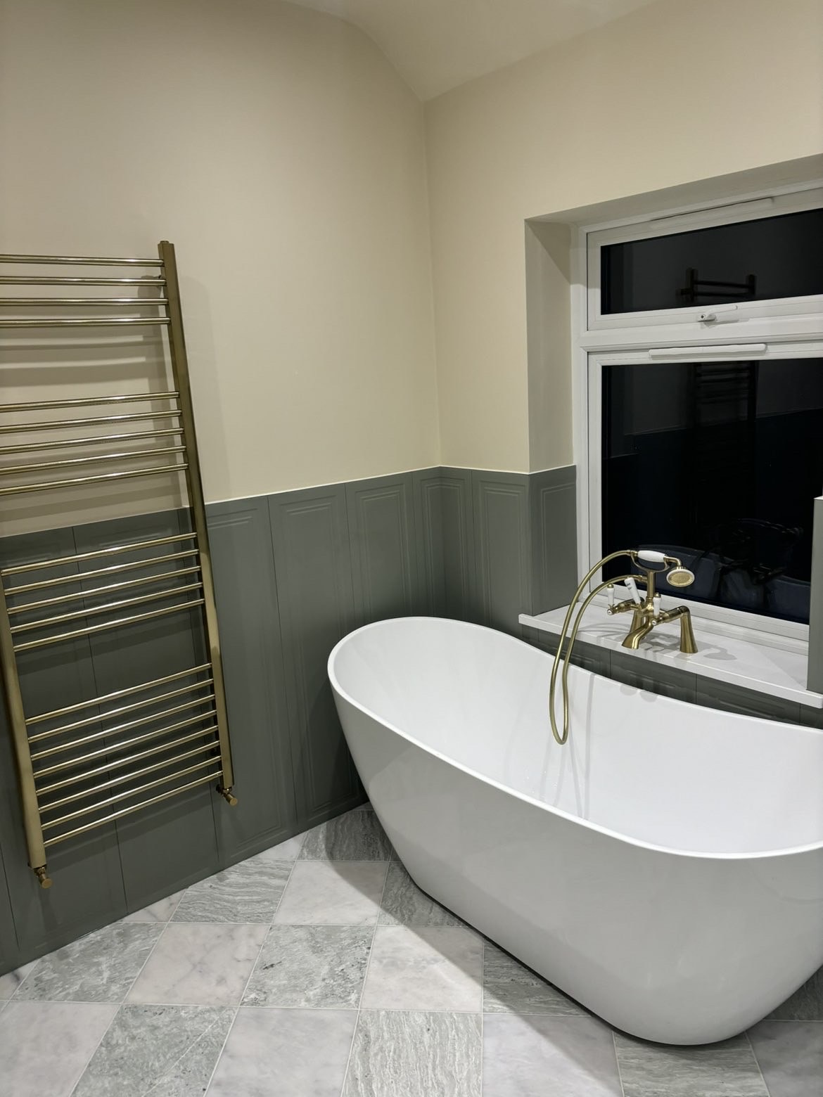 Brushed brass ladder heated towel rail on green panelled wainscoting beside the freestanding bath